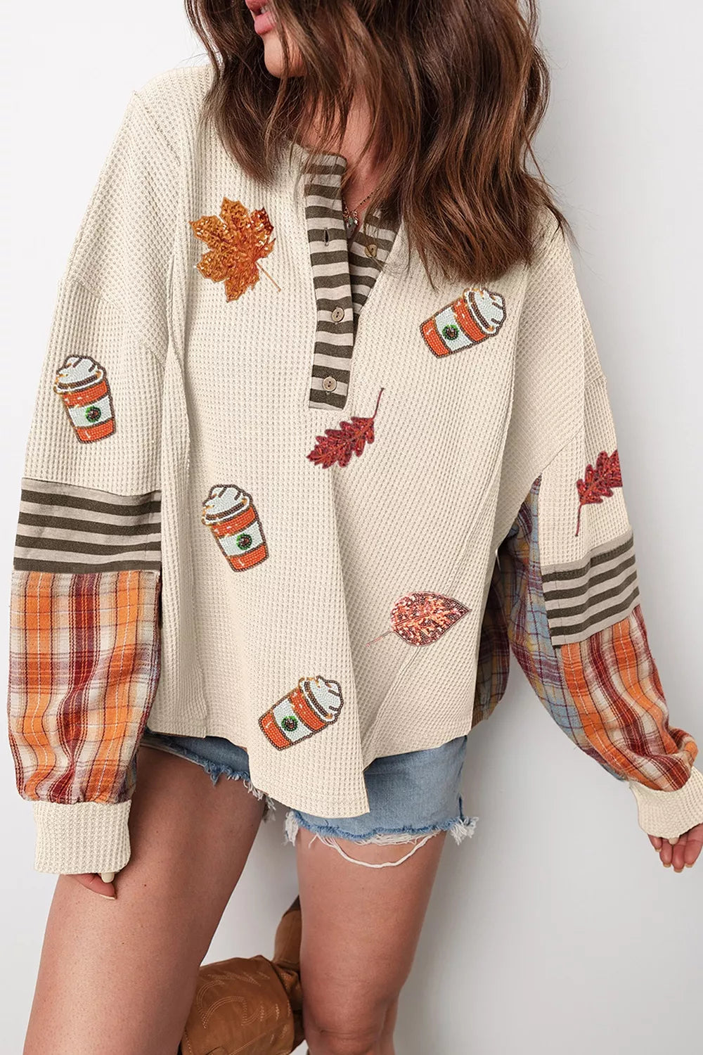 Woman wearing a cream waffle knit top with sequin autumn leaves and coffee cups, a striped button neckline, and orange plaid sleeve panels
