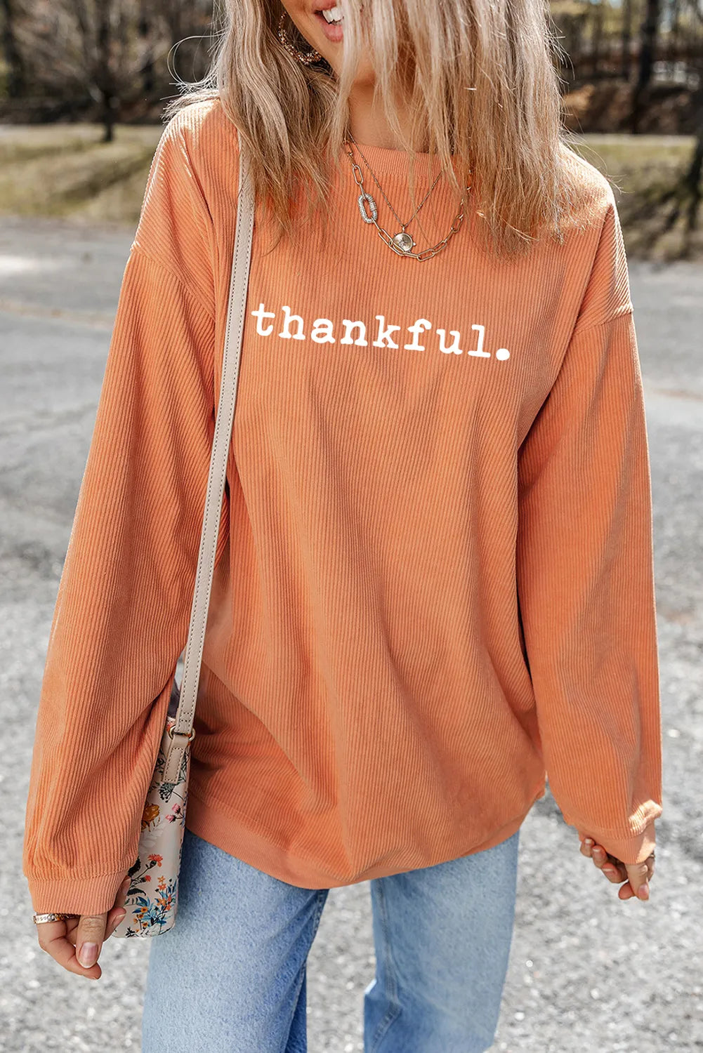 Woman wearing a pumpkin-orange ribbed sweatshirt with white “thankful.” lettering, styled with light-wash jeans