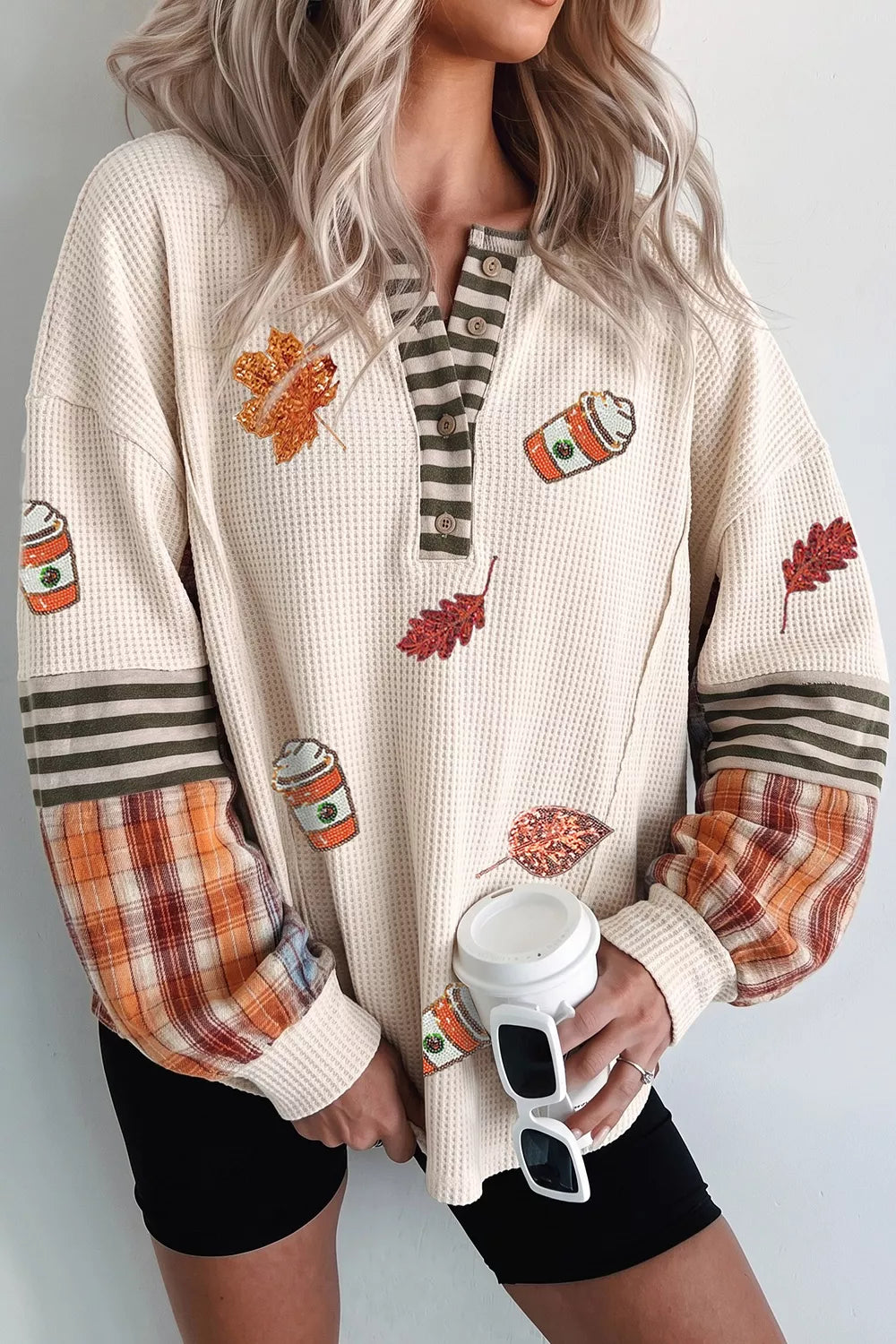 Woman wearing a cream waffle knit top with sequin autumn leaves and coffee cups, a striped button neckline, and orange plaid sleeve panels