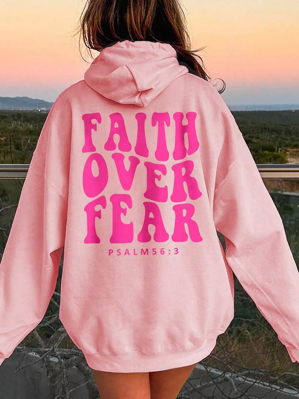 Person wearing a pink hoodie with 'Faith Over Fear' text outdoors.