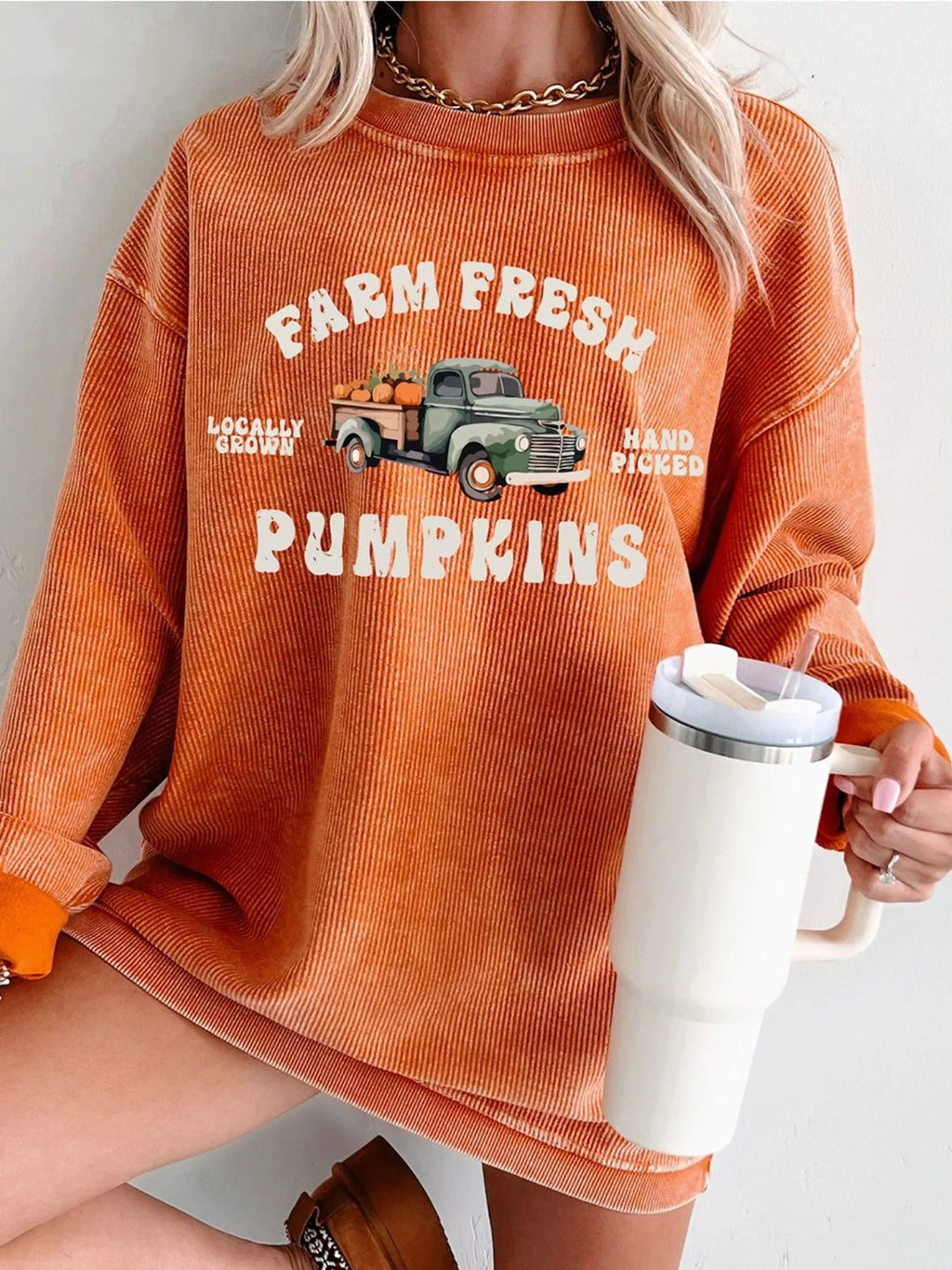 Woman wearing an orange ribbed sweatshirt with “Farm Fresh Pumpkins” lettering and a vintage green truck carrying pumpkins, paired with white jeans