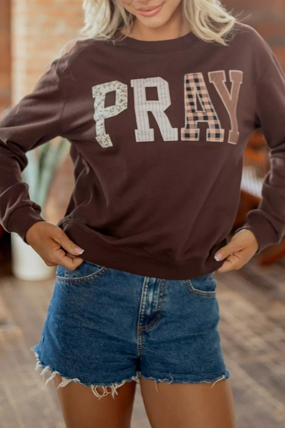 Woman wearing a chocolate brown crewneck sweatshirt with “PRAY” in large floral, striped, plaid, and solid neutral letters, paired with denim shorts