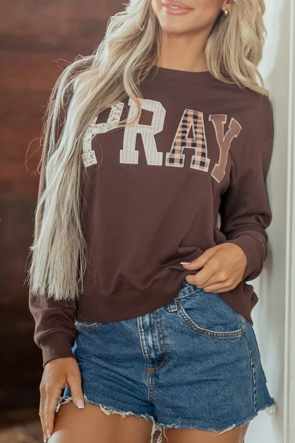 Woman wearing a chocolate brown crewneck sweatshirt with “PRAY” in large floral, striped, plaid, and solid neutral letters, paired with denim shorts