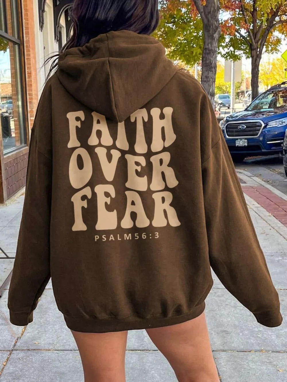 Person wearing a brown hoodie with 'Faith Over Fear' text on the back, standing on a sidewalk.