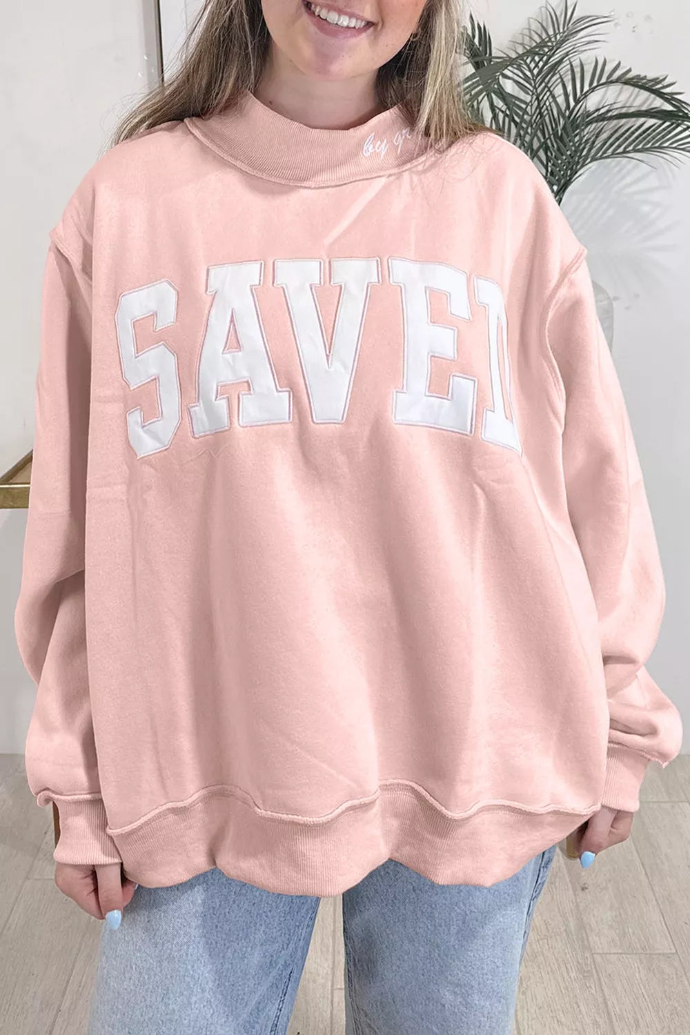 A person wearing an oversized dusty pink sweatshirt with the word 'SAVED' in large white block letters across the chest, paired with light blue jeans.