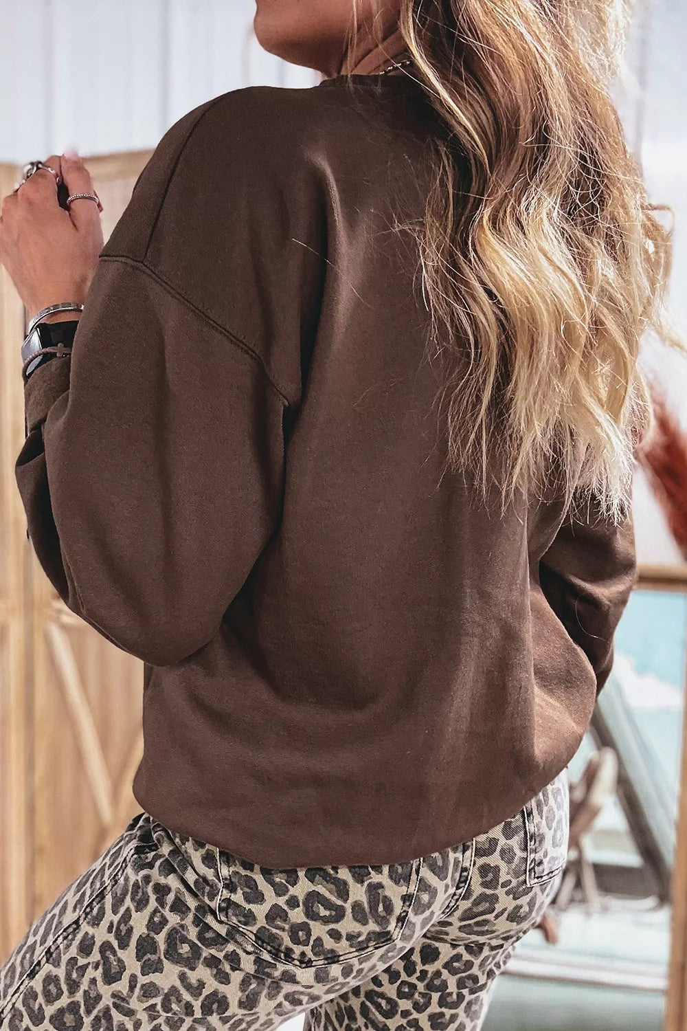 Woman wearing a chocolate brown crewneck sweatshirt with “PRAY” in large floral, striped, plaid, and solid neutral letters. 