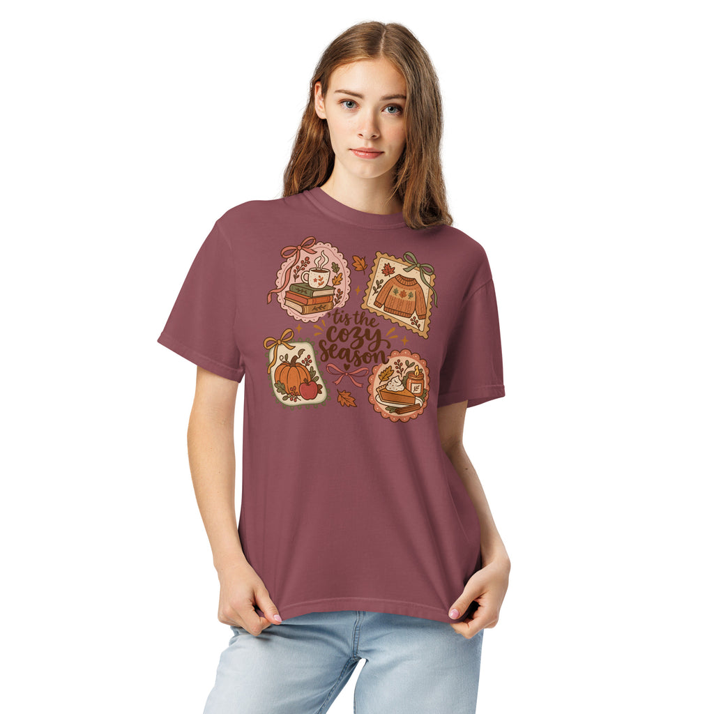 Model wearing a muted maroon “’Tis the Cozy Season” graphic tee with fall illustrations of books, a steaming mug, a sweater, pumpkins, and pie.