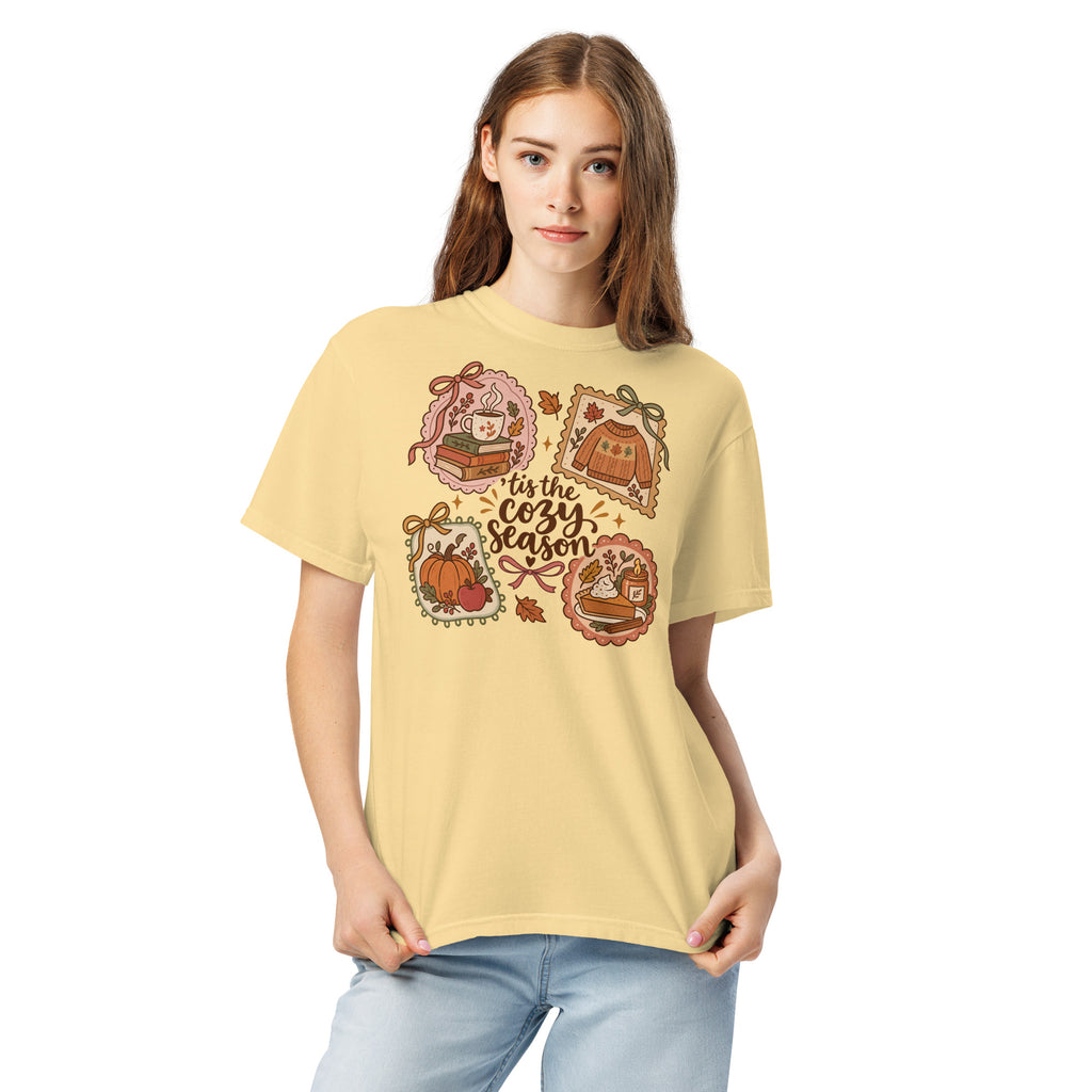 Model wearing a muted yellow “’Tis the Cozy Season” graphic tee with fall illustrations of books, a steaming mug, a sweater, pumpkins, and pie.