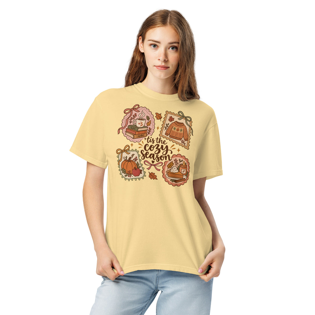 Model wearing a muted yellow “’Tis the Cozy Season” graphic tee with fall illustrations of books, a steaming mug, a sweater, pumpkins, and pie.