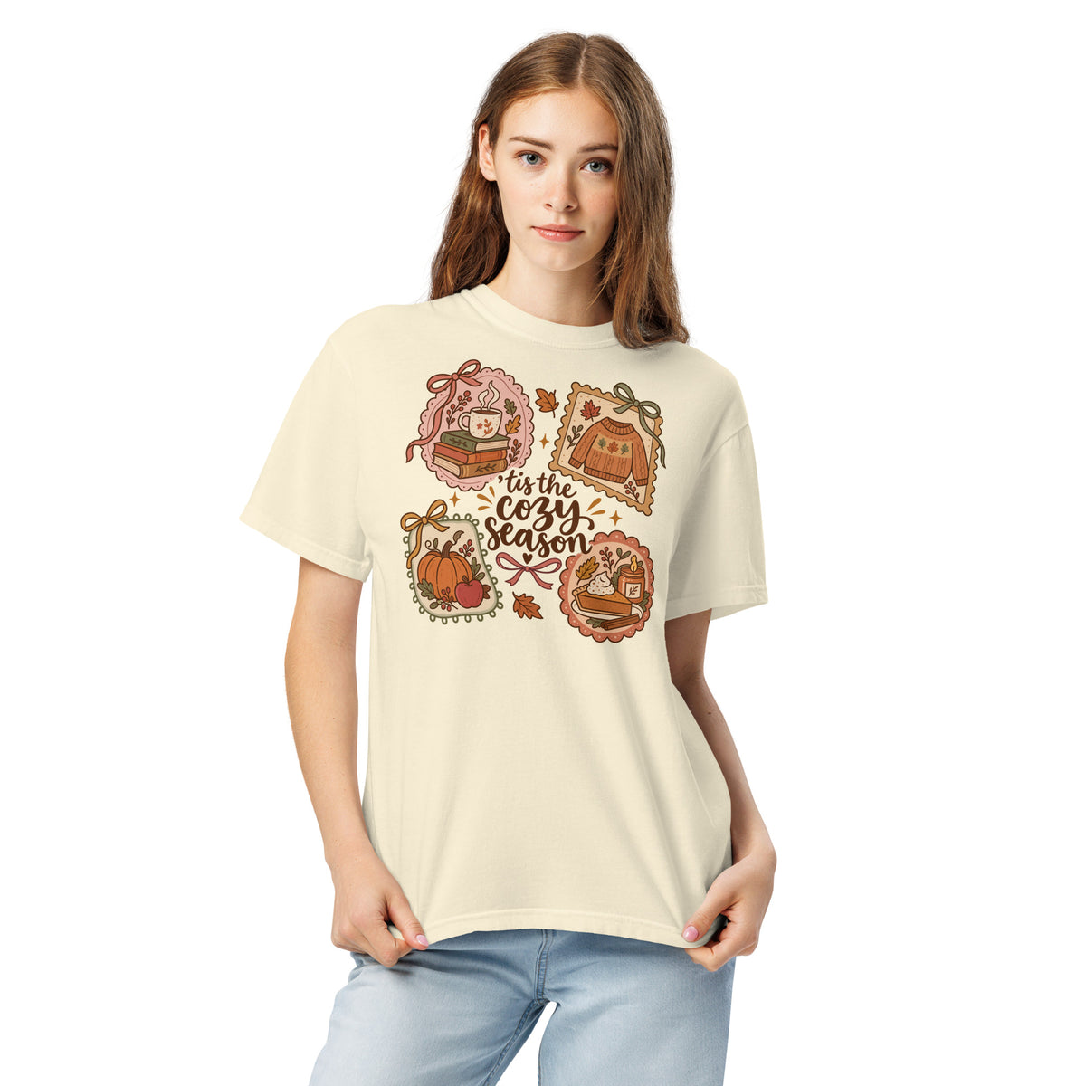 Model wearing a beige “’Tis the Cozy Season” graphic tee with fall illustrations of books, a steaming mug, a sweater, pumpkins, and pie.