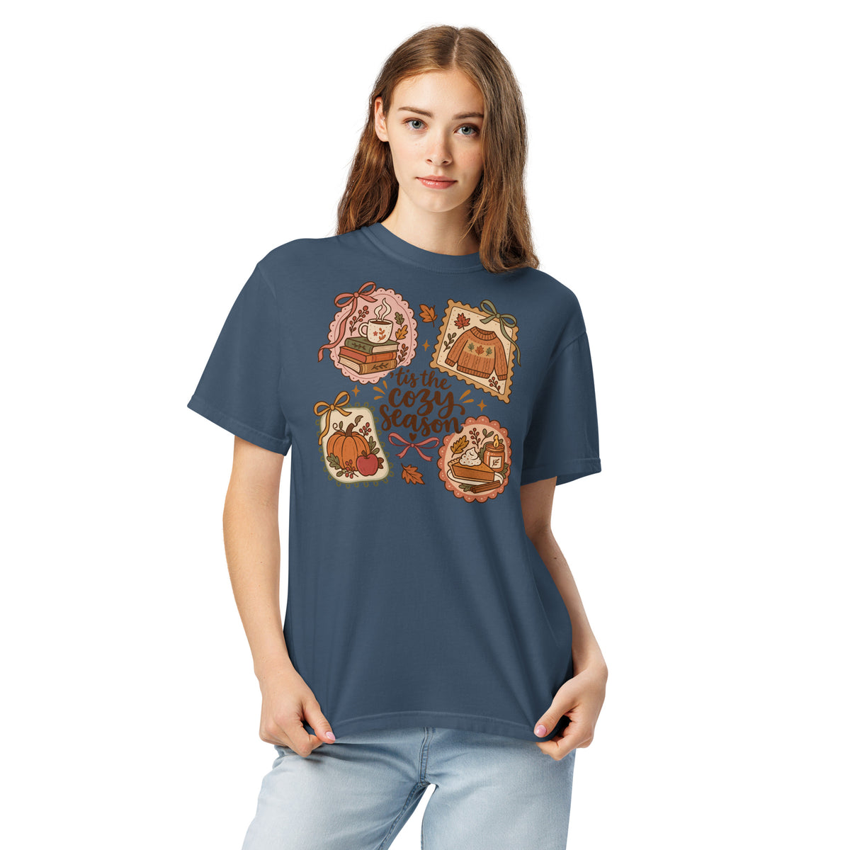 Model wearing a muted navy blue “’Tis the Cozy Season” graphic tee with fall illustrations of books, a steaming mug, a sweater, pumpkins, and pie.