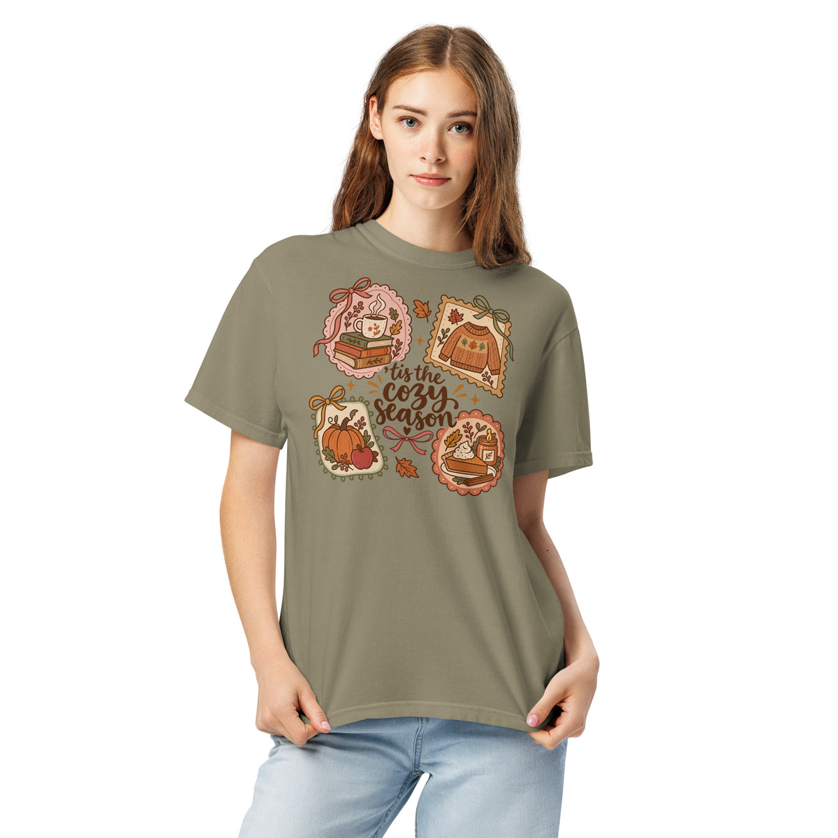 Model wearing a muted green “’Tis the Cozy Season” graphic tee with fall illustrations of books, a steaming mug, a sweater, pumpkins, and pie.
