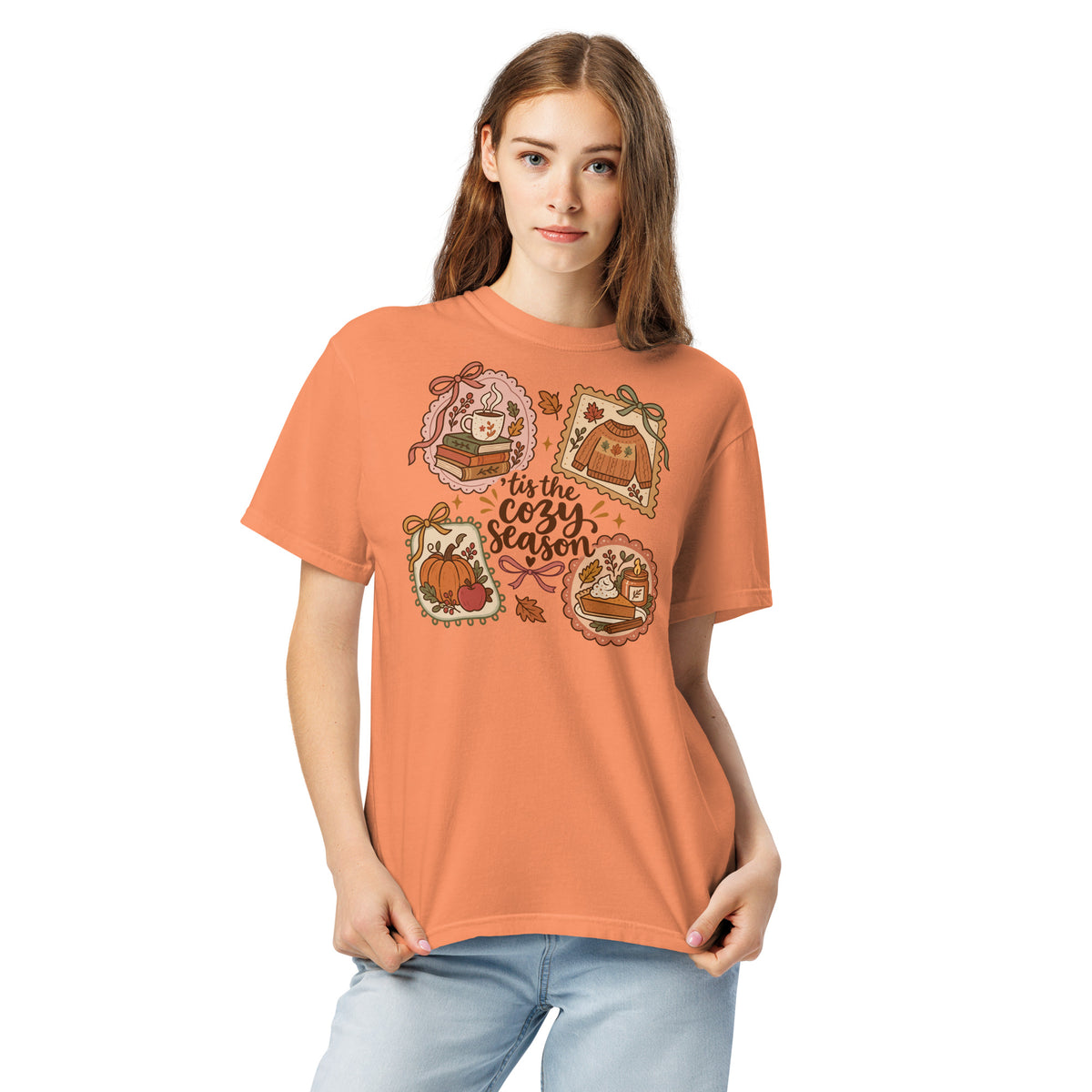 Model wearing an orange “’Tis the Cozy Season” graphic tee with fall illustrations of books, a steaming mug, a sweater, pumpkins, and pie.