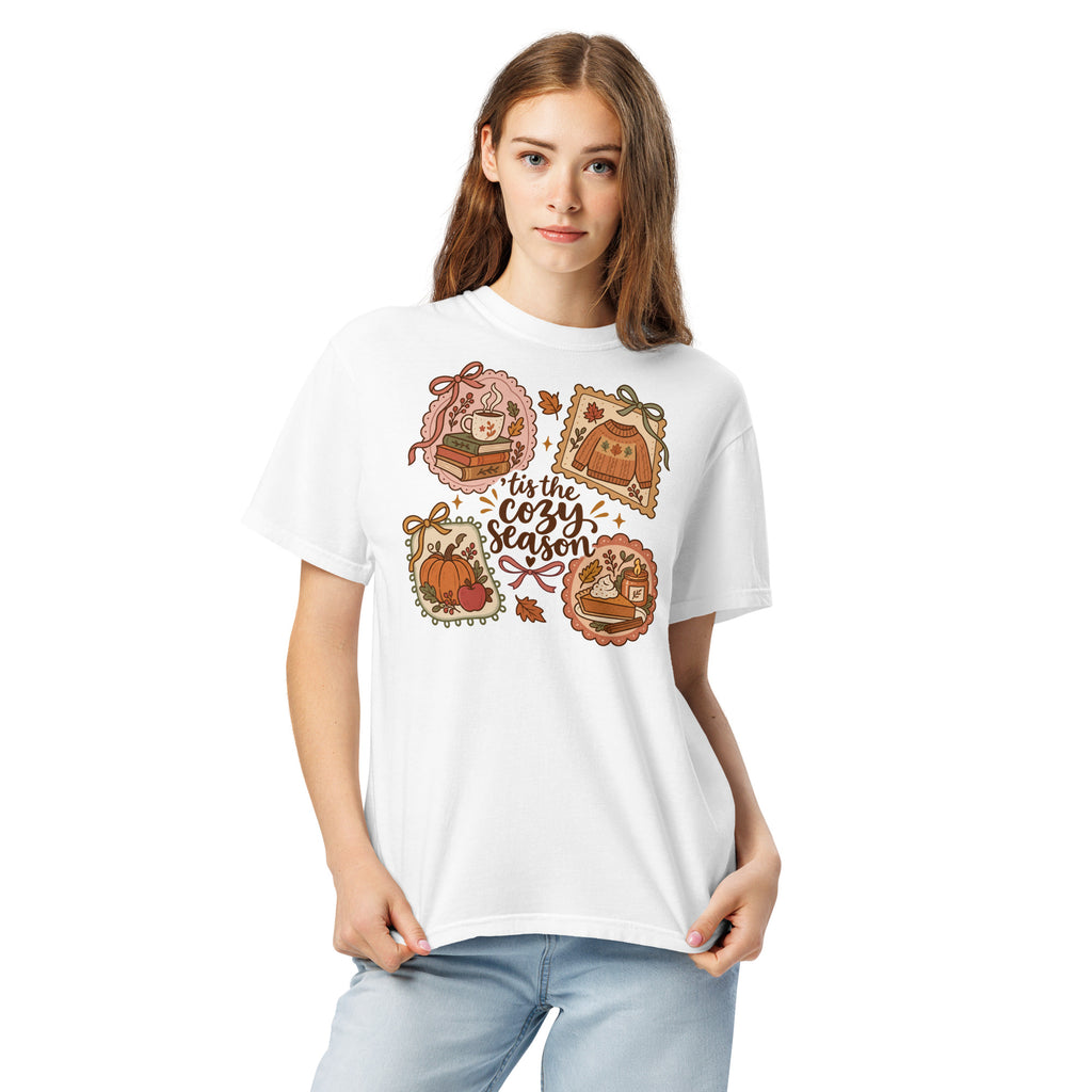 Model wearing a white “’Tis the Cozy Season” graphic tee with fall illustrations of books, a steaming mug, a sweater, pumpkins, and pie.