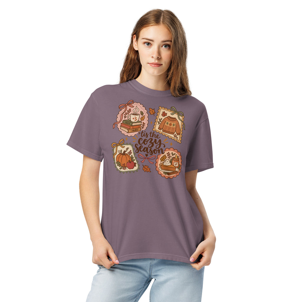 Model wearing a muted purple “’Tis the Cozy Season” graphic tee with fall illustrations of books, a steaming mug, a sweater, pumpkins, and pie.