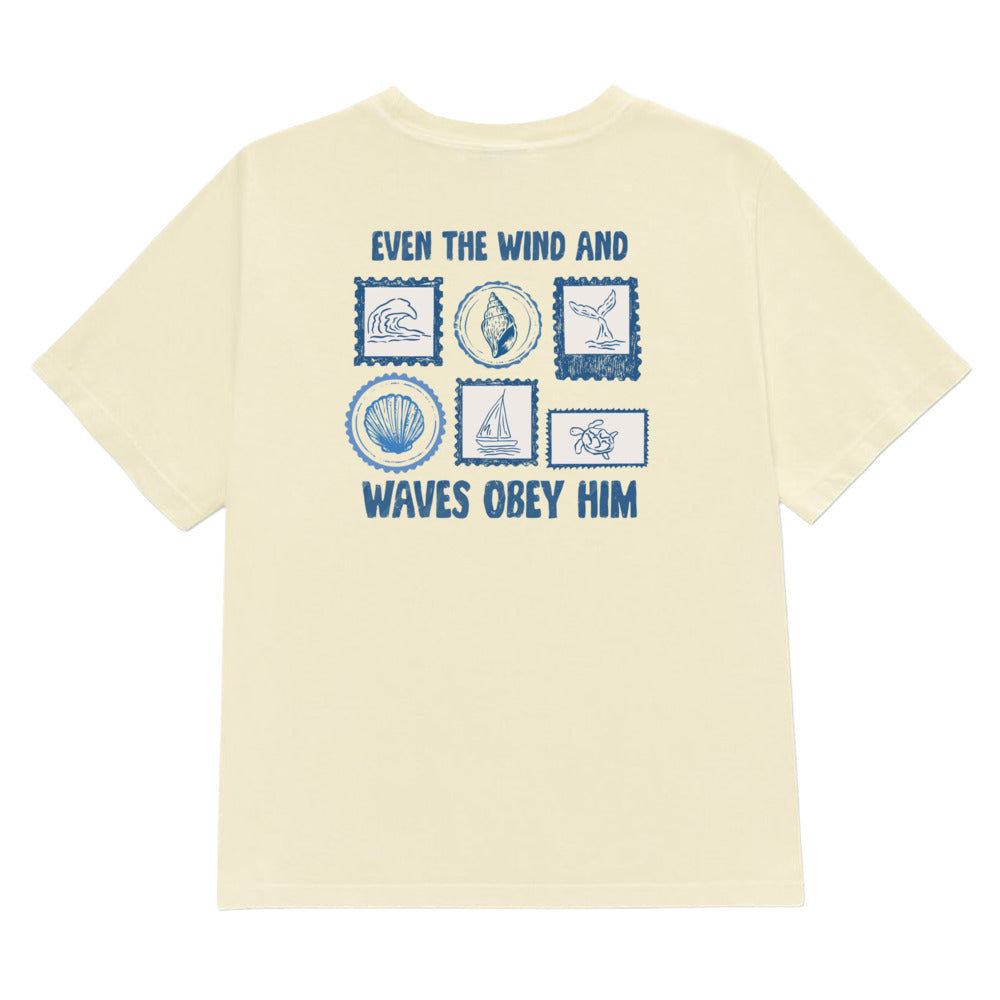 A butter yellow short-sleeve t-shirt with a blue graphic print on the back. The print features the text 'EVEN THE WIND AND WAVES OBEY HIM' surrounding a grid of six blue postage stamp-like illustrations depicting a wave, a seashell, a sailboat, a wave breaking, a seashell, and a wave.